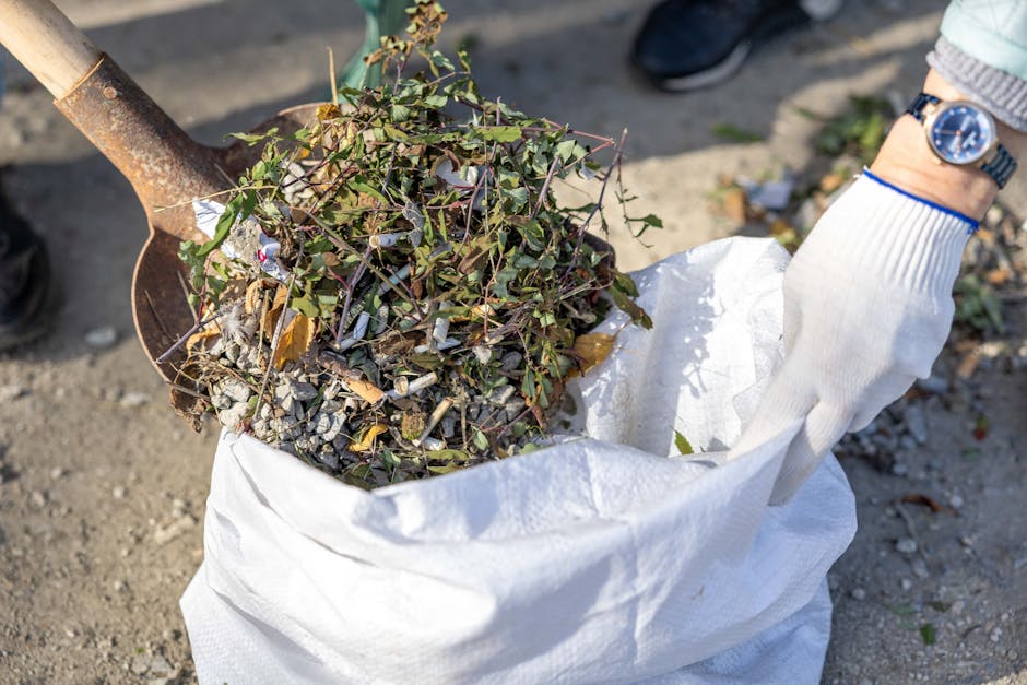A close-up view of a person wearing white gardening gloves holding a large white sack filled with garden waste, including a mix of chopped plant material, small branches, and leaves. The background sh