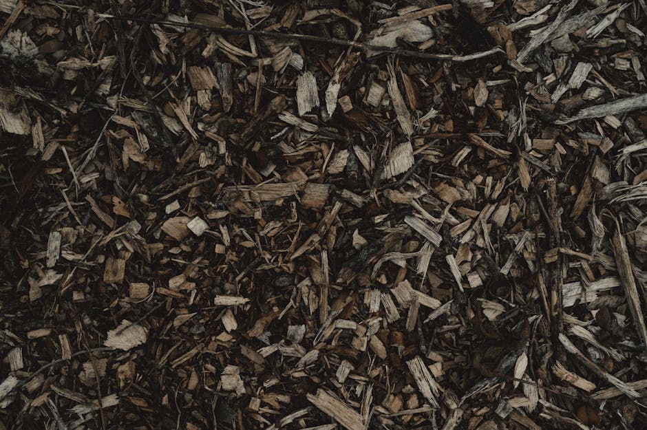 A close-up view of a person wearing white gardening gloves holding a large white sack filled with garden waste, including a mix of chopped plant material, small branches, and leaves. The background sh